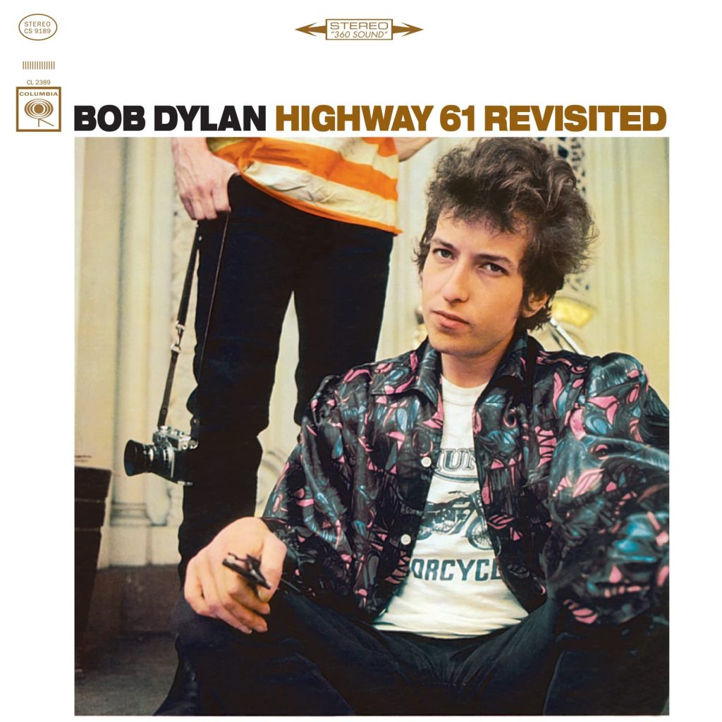 The album cover for Highway 61 Revisited by Bob Dylan. A white background with a photo of Bob Dylan, a pale-skinned man with disheveled brown hair, seated and wearing a multicolored button up short over a white graphic tee. Behind him is another person in black pants and an orange and white striped shirt carrying a film camera dangling from a strap.
