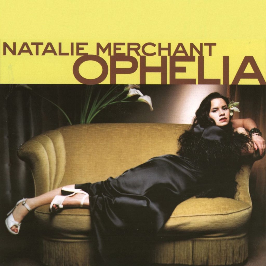 The album cover for Ophelia by Natalie Merchant. Natalie Merchant, a pale-skinned woman with black hair and a white lily behind her ear wears a black satin dress and white high heels and lounges on a golden sofa with a fringed bottom. Behind her is a potted white lily and brown curtains. The top of the album is a yellow rectangle with the title in brown text.