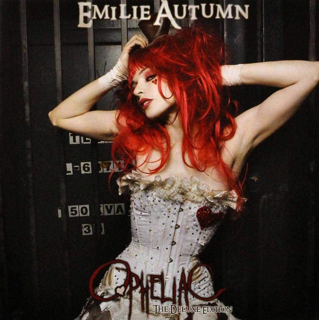 The album cover for the deluxe edition of Opheliac by Emilie Autumn. Emilie Autumn, a young woman with pale skin and wild red hair wears a white, sleeveless vintage corset dress with a glittering red heart on the left side of the chest. There are other lace and jewel embellishments on the corset. The woman has both of her arms raised behind her head and a red heart drawn underneath her left eye. She stands in front of a black door.
