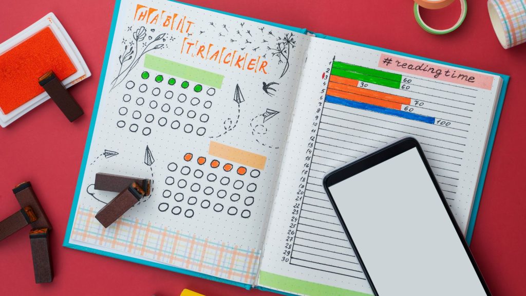 Open journal with a habit tracker on the left, featuring doodles and colorful circles. Right page shows a reading log. A smartphone lies on one page.