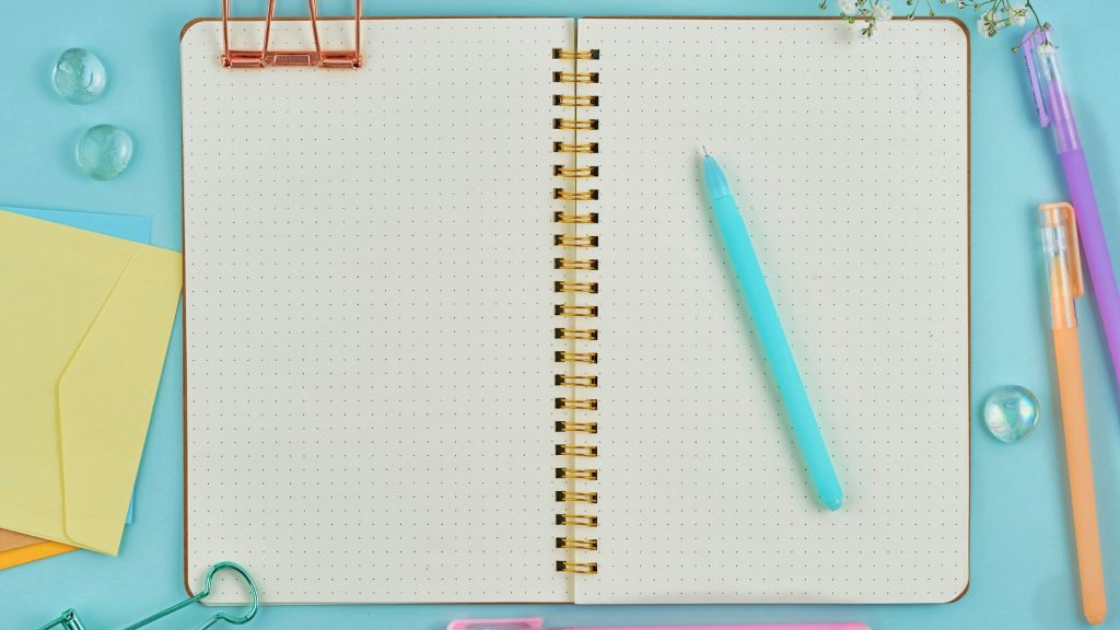 Open spiral notebook with dotted pages on a light blue surface, surrounded by pastel pens, paper clips, and envelopes. A turquoise pen rests on the notebook.