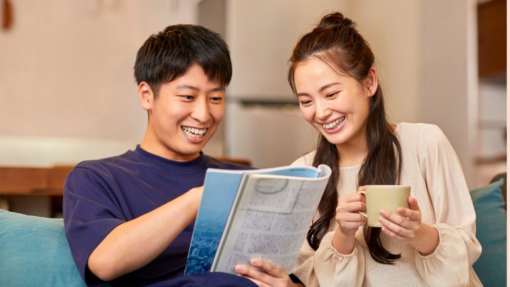 Two people sit on a sofa, smiling and engaged while reading a magazine. The woman holds a coffee mug, creating a warm, cozy, and cheerful atmosphere.