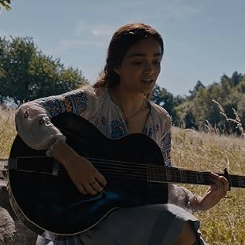 Movie still from The Ballad of Songbirds and Snakes, Lucy Gray Baird playing the guitar in the meadow.