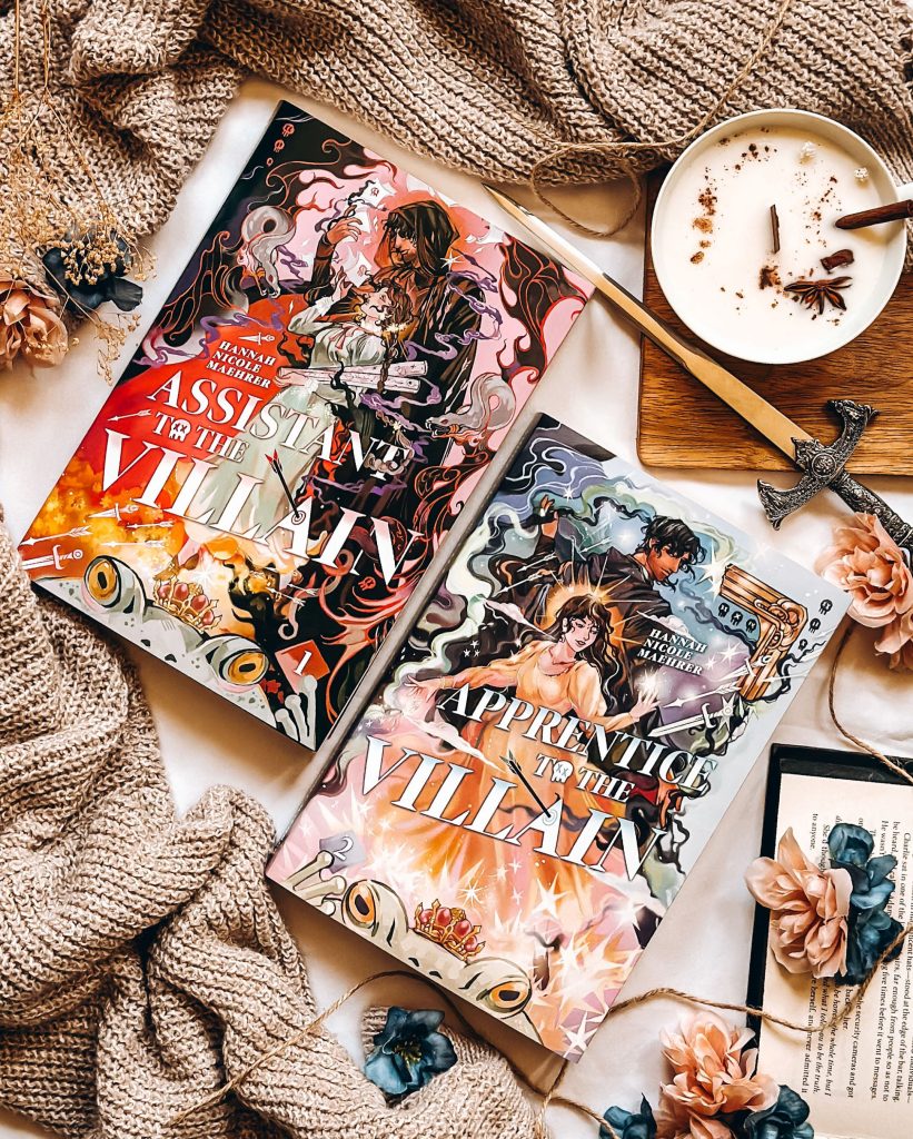 Flatlay created by Bookstagrammer Britt of her favorite book covers, the Fox and Wit special edition of Assistant to the Villain and Apprentice to the Villain. The books are surrounded by pale blue and pink flowers, a dagger, a candle filled with cinnamon sticks and spice, a beige sweater, and an open book in the corner.
