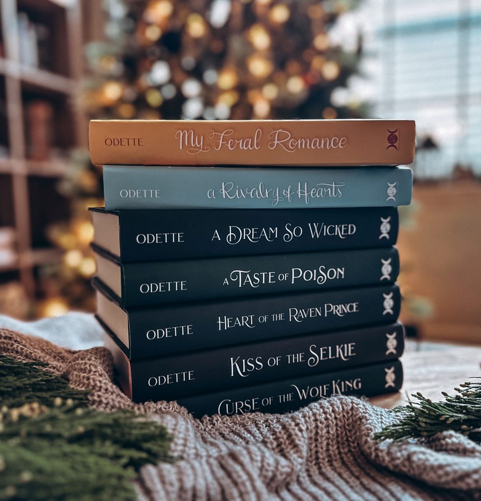Book stack curated by Bookstagrammer Britt of her collection of one of her favorite author, Tessa Odette's novels. Novels Include Curse of the Wolf King and Rivalry of Hearts with the books navy blue, light blue, and tan spines facing the camera laid on a sweater with pine leaves in the corners of the frame and a blurred Christmas tree in the background.