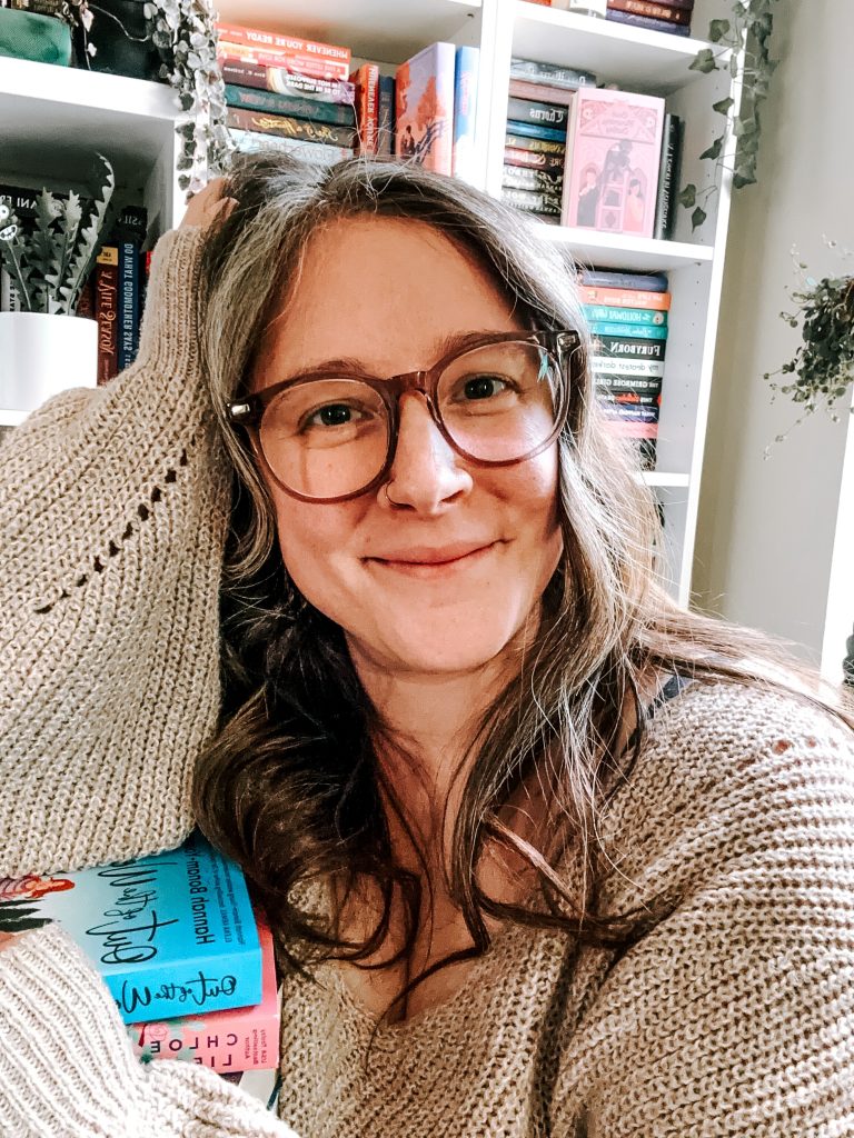 A selfie of Bookstagrammer Britt in her home library with a background of book filled shelves and her arm resting on a stack of books while she's wearing a beige sweater and brown glasses.