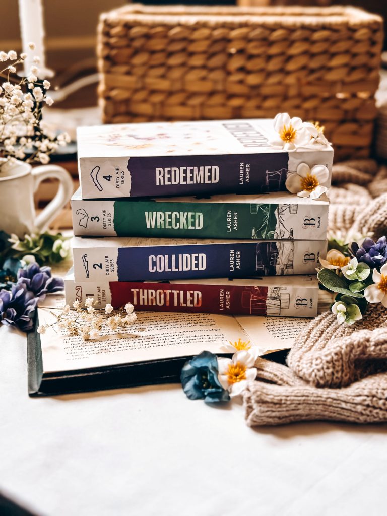 Book stack created by Bookstagrammer Britt of Lauren Asher's Dirty Air Series, including the titles Redeemed, Wrecked, Collided, and Throttled, with the spines facing the camera. Britt laid the book stack laid on an open book with sprayed black edges, and decorated the scene with various flowers, a sweater, and a wicker basket in the background.