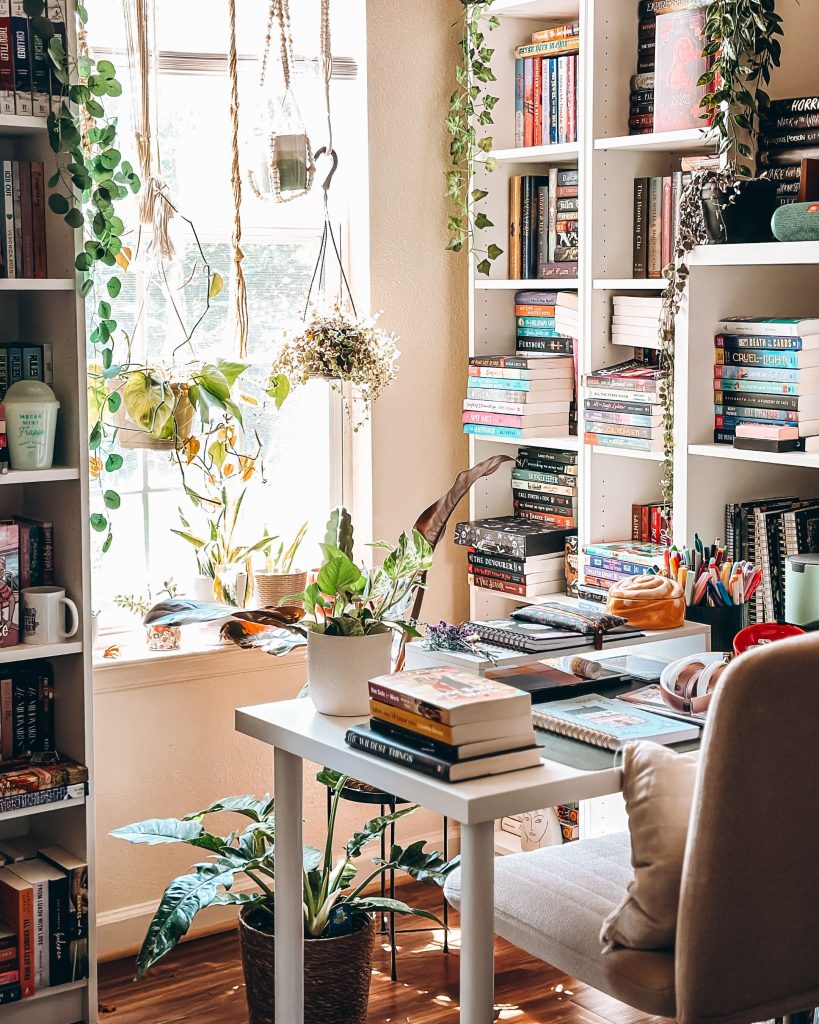 Bookstagrammer Britt's desk space and home library of neat, white bookshelves filled with books going in vertical and horizontal directions. Her desk is filled with books and is facing a window that has sunlight pouring through it, illuminating the many hanging plants and vines in the window.