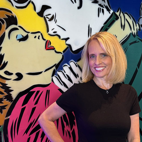 Romance Author Lauren Blakely standing before a wall with a painting of two lovers embracing as they lean in to kiss.