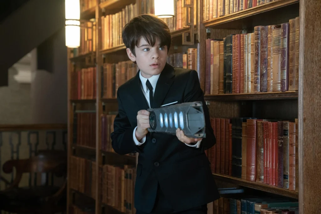 A young boy, Artemis Fowl, in a black suit holds a futuristic device in a dimly lit library. He looks alert, surrounded by shelves of old, leather-bound books.