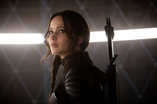 Jennifer Lawrence, as Katniss Everdeen in The Hunger Games, with braided hair stands in a dark, futuristic setting, looking to the side with determination. She wears a black outfit and carries a bow and arrows.