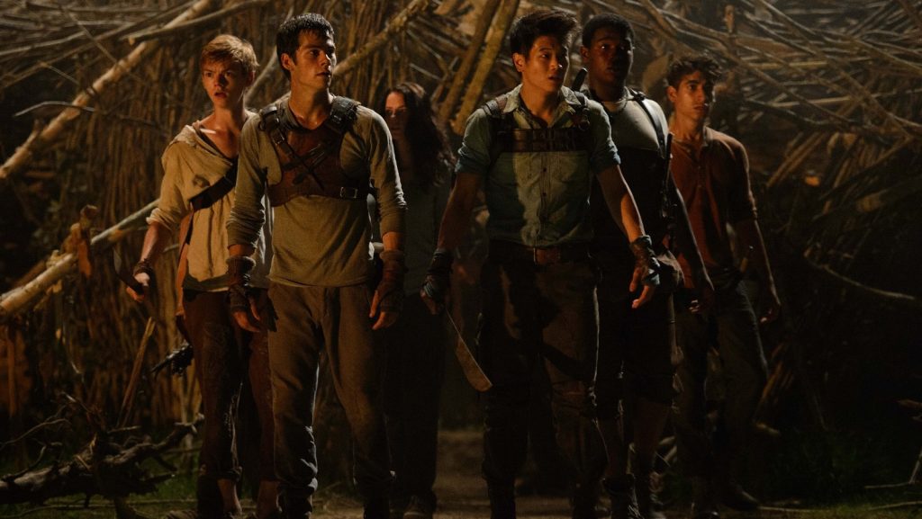 A group of six young boys, from The Maze Runner movie, stands in the shadows of a forest. They wear rugged clothing and carry weapons, exuding determination and teamwork.