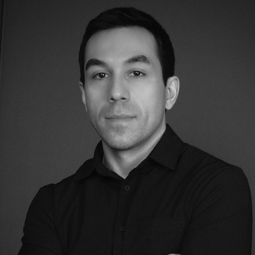 Nick Medina, a light-skinned man with short black hair and a black collared shirt, looks at the viewer with a neutral expression on his face; the image is in black and white with a solid background.