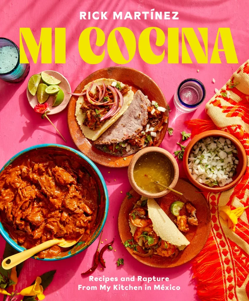 Mi Cocina: Recipes and Rapture from My Kitchen in Mexico by Rick Martinez features a pink table with plates of food and a drink. The plates have rice, a stew, tacos, salsa, limes, and a tablecloth with a traditional design on the edge of the cover.