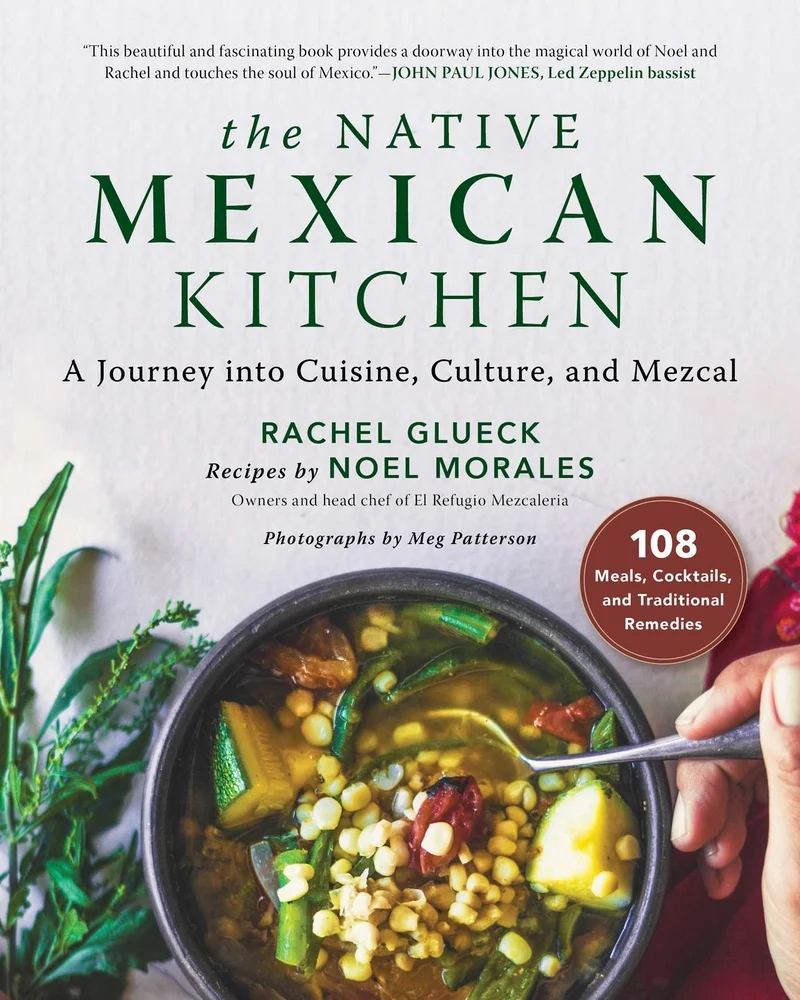 The Native Mexican Kitchen: A Journey into Cuisine, Culture, and Mezcal by Rachel Glueck and Noel Morales depicts a table with a bowl of Mexican soup with corn, zucchini, and chiles, and a hand with a spoon in the bowl. Next to the bowl are herbs with the title above it.