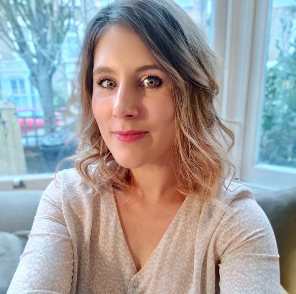 Author Katherine Faulkner headshot as she sits on a plush couch, holding the camera up as a selfie and looks directly into the camera. She wears a cream, off-white colored blouse and has her blonde hair in loose curls down that frame her face. The background is a window that looks out onto a busy street behind her.