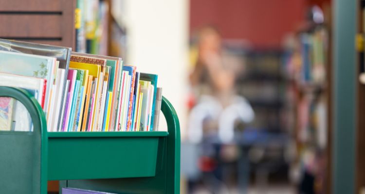 A green cart filled with colorful books against a blurred library.