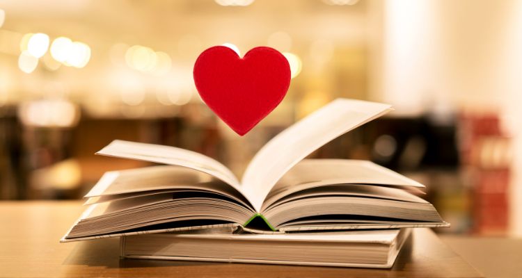 An open book with a red heart above it. There is a closed book under it that seems to be resting on a wooden surface.