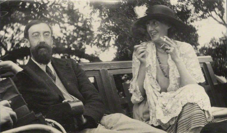 A photo taken in June 1923 of Lytton Strachey and Virginia Woolf sitting on a bench. Lytton is wearing a three-piece suit, and Virginia is wearing a lace cover and a large, wide-brimmed hat with a giant feather on the top. Virginia is smoking a cigarette and laughing.
