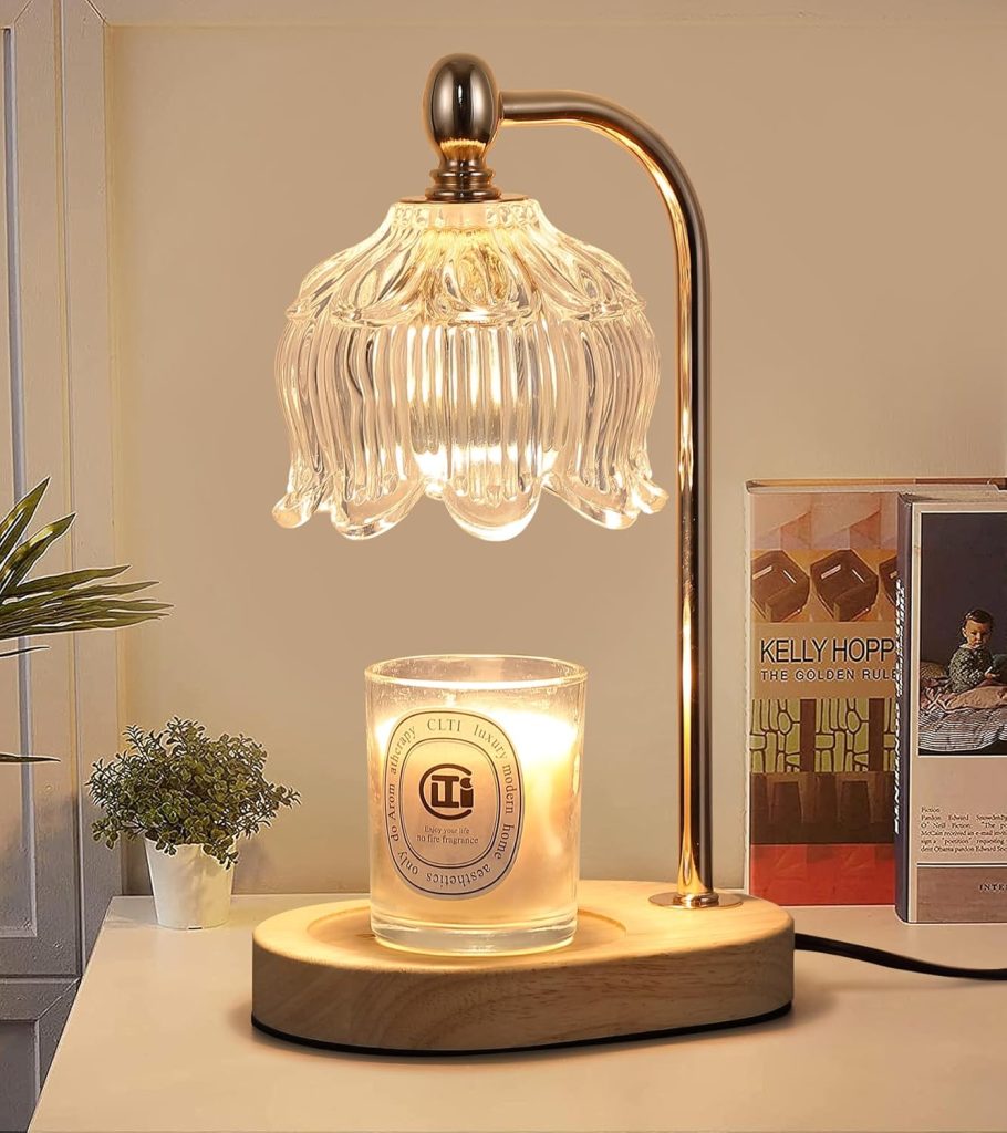 The image features a lit glass flower head candle warmer with a candle on the base. In the background are books, and a plant is placed upon a table top.