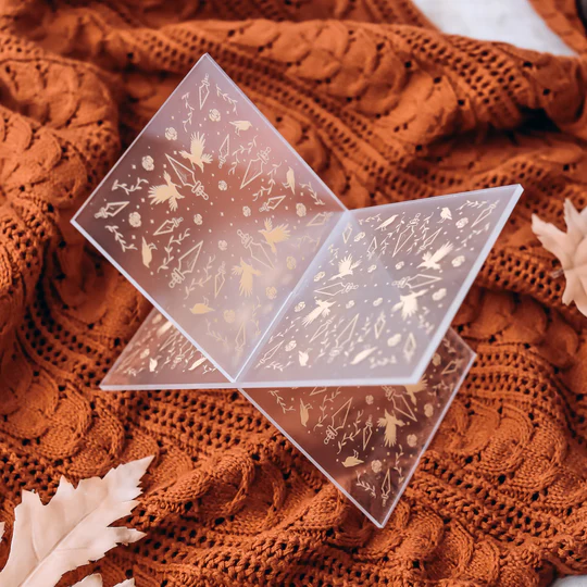 The image features a plastic X book holder with dagger heads, dragons, vines, and leaves. Under the holder is a knit blanket with leaves.