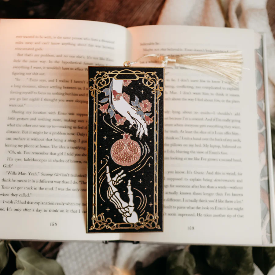 This image depicts an open book with a bookmark featuring two hands wrapped in vines, reading towards a pomegranate. One of the hands is a skeleton, and one is normal.