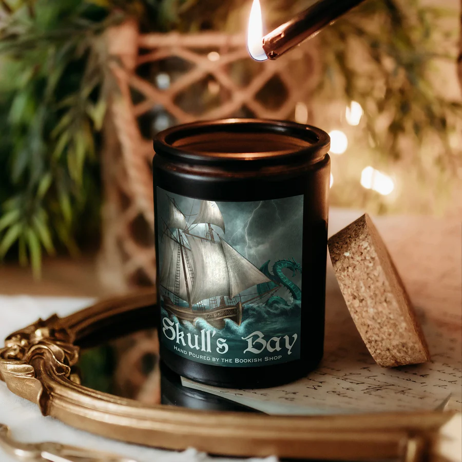 The image features a black candle placed on a mirror with a pirate ship in the ocean, with the words Skull's Bay underneath. Next to the candle is a cork, and on top is a lighter with a flame. In the background, there is greenery and a woven basket.