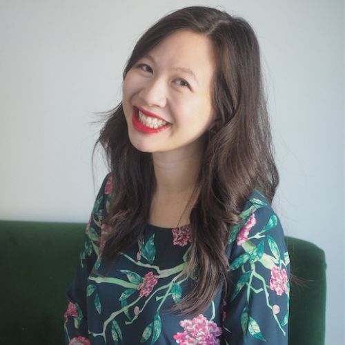Headshot of author Elizabeth Lim. She is wearing a floral print, quarter-sleeve top and is seated on a green couch. She is smiling with teeth and has her head slightly tilted to the right side.