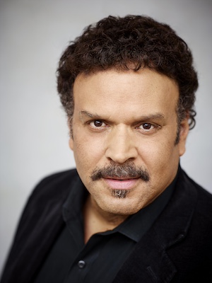 Headshot of YA author Neal Shusterman. He has slightly curly brown hair and facial hair that is well-groomed. He is wearing a black, collared buttoned down shirt and faces the viewer. He has a pensive look on his face.