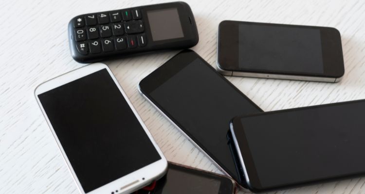 A collection of mobile devices arranged on a light wooden surface. There are six devices total: one basic cell phone with a physical keypad and small screen at the top, and five smartphones of various sizes with dark screens. The smartphones include both black and white/silver colored models, and they appear to be different makes and models lying flat on the wooden table or desk surface.