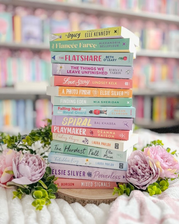 Tall stack of romance and contemporary fiction books displayed on a wooden round, featuring titles including The Legacy by Elle Kennedy, The Flatshare by Beth O'Leary, Love Story by Lindsey Kelk, and Powerless by Elsie Silver, decorated with pink peonies and green foliage against a blurred pastel bookshelf background.