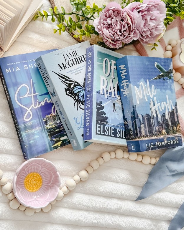 A styled Bookstagram flat lay featuring four romance novels with predominantly blue and teal covers arranged on a white textured fabric background. The visible titles include "Stir" by Mia Sheridan, a Jamie McGuire book, "Of Rage and Ruin" by Elsie Silver, and "Make Me High" by Liz Tomforde. The books are artfully surrounded by aesthetic props including soft pink peonies, fresh green eucalyptus sprigs, natural wooden beads arranged in curved lines, and a small pink ceramic bowl with a yellow center.