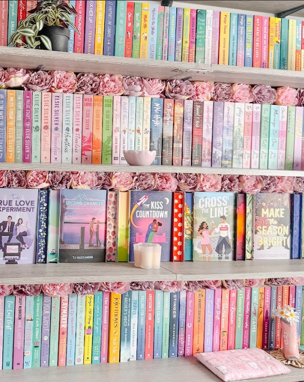 A detailed view of an extensively organized bookshelf featuring four shelves of color-coordinated books decorated with pink paper flower garlands along each shelf edge. The top shelf displays books arranged by color in a rainbow pattern with a small potted plant. The second shelf continues the color coordination theme with books arranged from darker to lighter spines, including Elsie Silver titles, with a white ceramic bowl as decoration. The third shelf showcases several face-out books including "True Love Experiment," "Second Chance," "Kiss Countdown," "Cross the Line," and "Make Me Season," along with white pillar candles for styling. The bottom shelf features more color-coordinated books ranging from blues and greens to pinks and corals, with what appears to be a pink notebook or planner at the end.