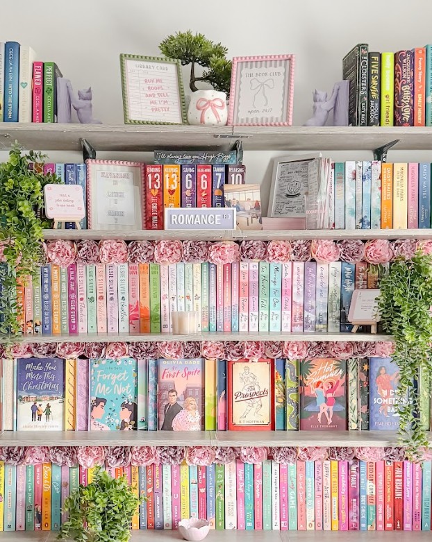 An organized bookshelf display featuring five shelves of color-coordinated books with feminine Bookstagram styling. The top shelf contains books with decorative quotes cards, small potted plants, and purple bookends shaped like cats. The second shelf features a prominent white "ROMANCE" sign among various books and decorative elements. The middle shelf showcases books arranged by color with pink paper flower garlands along the edge, creating a rainbow effect of pastels and bright colors. The fourth shelf displays face-out books including titles like "Beach This Christmas," "People We Meet on Vacation," and other contemporary romance novels, also decorated with pink floral garlands. The bottom shelf continues the color-coordinated theme with books arranged by spine color from pink to purple to blue. Throughout all shelves, small green plants, decorative accessories, and careful styling create the signature girly, organized aesthetic typical of Bookstagram content.