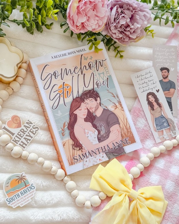 A Bookstagram flat lay featuring "Somehow Still You" by Samantha Anne, a contemporary romance novel with an illustrated cover showing a couple embracing. The book is accompanied by a matching character bookmark and surrounded by soft pink peonies, fresh greenery, and white wooden beads arranged decoratively. A bright yellow fabric bow and small enamel pins add pops of color to the white textured fabric background.