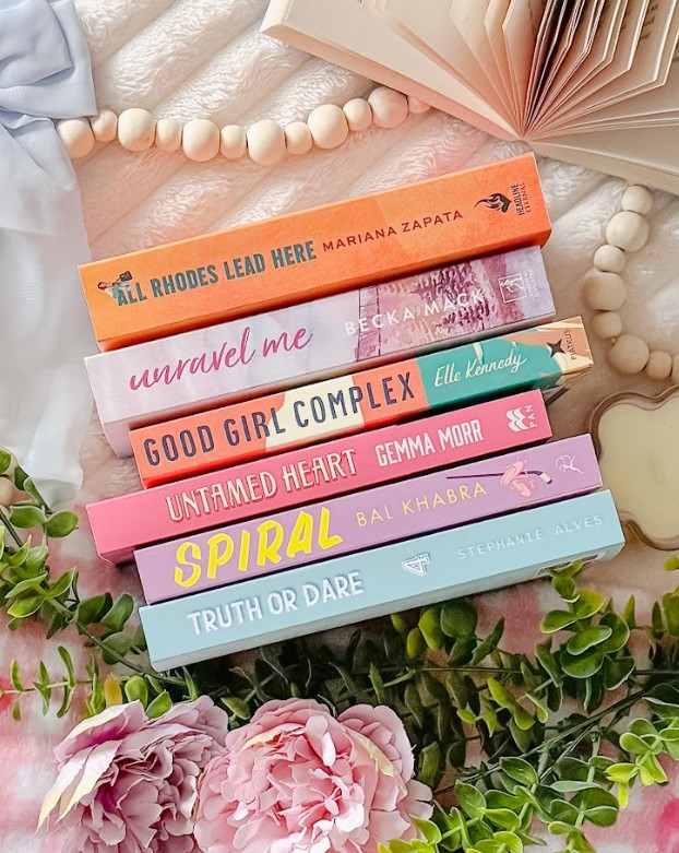 A carefully styled Bookstagram flat lay featuring a stack of romance and contemporary fiction books with colorful spines arranged vertically. The visible titles include All Rhodes Lead Here by Mariana Zapata (orange), Unravel Me by Becka Mack (pink), Good Girl Complex by Elle Kennedy (teal), Untamed Heart by Gemma Morr (pink), Spiral by Bal Khabra (purple with yellow text), and Truth or Dare by Stephanie Alves (light blue). The books are surrounded by aesthetic props, including soft pink peonies, green eucalyptus sprigs, natural wooden beads arranged in curves, white fabric, and an open book with fanned pages in the background.