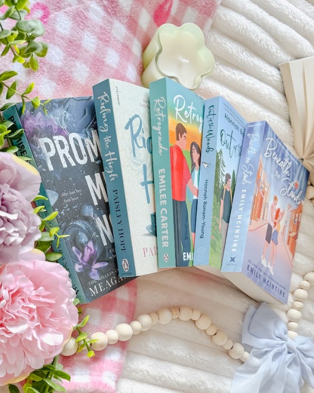 A Bookstagram flat lay featuring a collection of contemporary romance novels arranged in a fan pattern, including titles by authors like Emily Carter and others with colorful illustrated covers. The books are surrounded by soft pink peonies, fresh green foliage, and natural wooden beads arranged decoratively across white textured fabric and pink gingham. A small cream-colored flower-shaped ceramic bowl and light blue fabric bow add delicate styling touches.