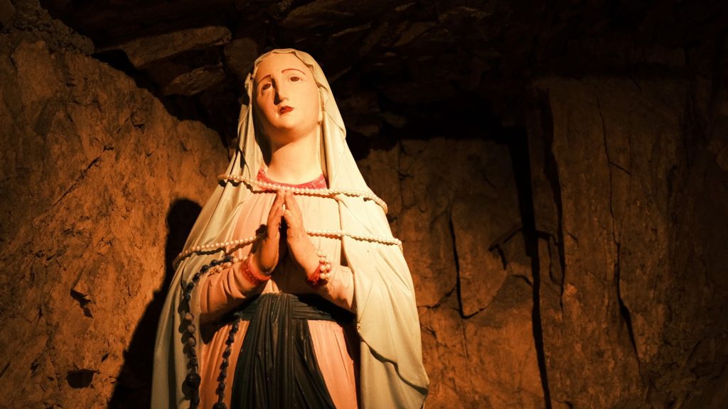 A statue of the Virgin Mary in a dimly lit cave-like setting. She looks up toward the sky with a sorrowful expression, her hands pressed together in prayer. A rosary hangs from her right arm, and pearls are strung around her body.
