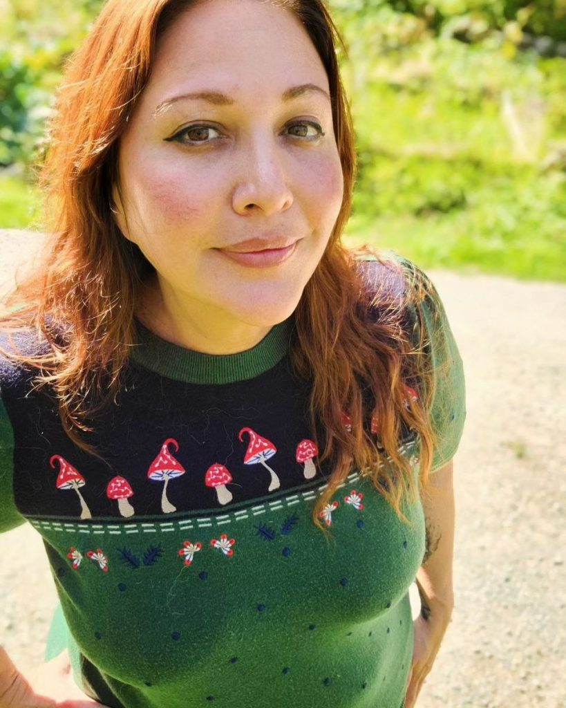 Headshot of Cassandra Newbould. She is looking up at the camera smiling, and is outside in a sweater. It is sunny out and she is on a concrete path with grass behind her.
