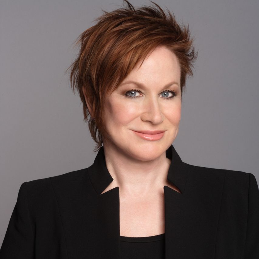 Christina Dodd, a light-skinned woman with short red hair, smiles at the viewer. She wears a black top and black blazer, and the background is gray.