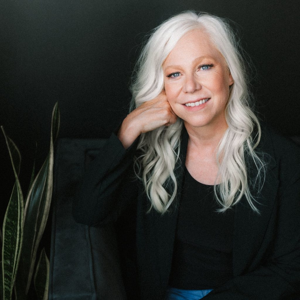 Heather Gudenkauf, a light-skinned woman with long white hair, rests her face on her right hand while smiling at the viewer. She wears a long-sleeved black shirt and denim jeans and sits next to a plant with long leaves. The background is black.