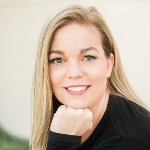 Samantha Downing, a light-skinned woman with long blonde hair, rests her chin on her fist while smiling at the viewer. She wears a long-sleeved black top, and the background is cream with green at the bottom.