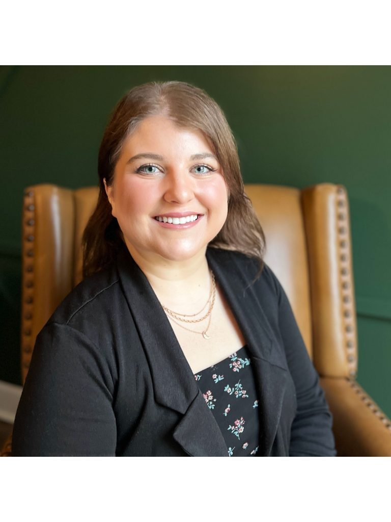 Kiersten Modglin, a light-skinned woman with light brown hair, sits in a brown armchair and smiles at the viewer. She wears a black blazer and black top with flowers on it, and the wall behind her is dark green.