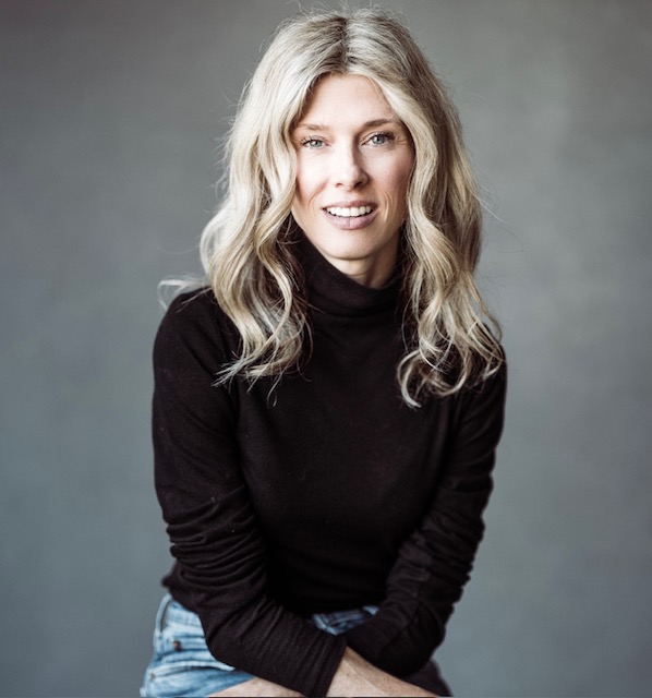 Ashley Audrain, a light-skinned woman with medium-length blonde hair, smiles at the viewer. She wears a long-sleeved black top and denim jeans, and the background is gray.