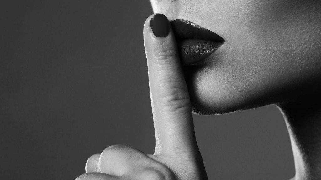A woman holding her finger to her lips in black and white; her lips and fingernail are painted.
