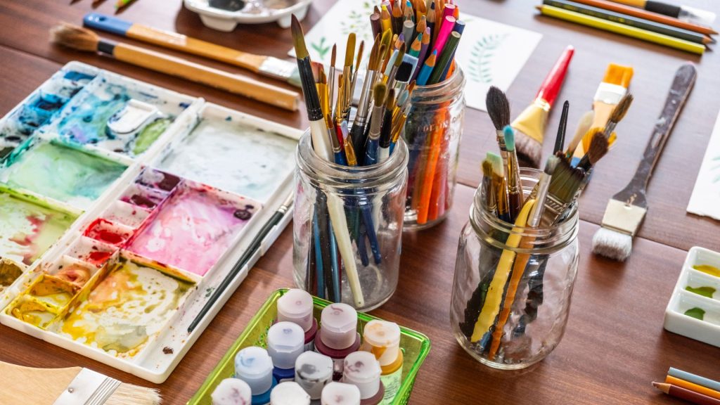 A vibrant art studio scene with jars of paintbrushes, a colorful watercolor palette, and small paint containers on a wooden table. The tools convey creativity.