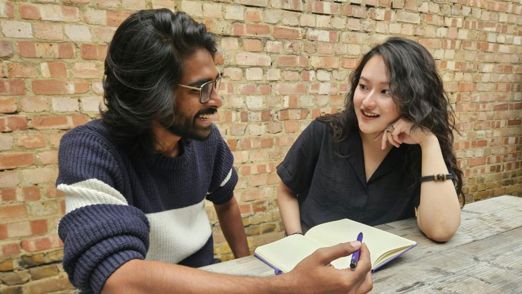 A man with brown skin, shoulder-length dark hair, a beard, and glasses holds a pen while talking to a light-skinned woman with long, wavy dark hair. They sit at a wooden table in front of a brick wall, a notebook lying open between them.