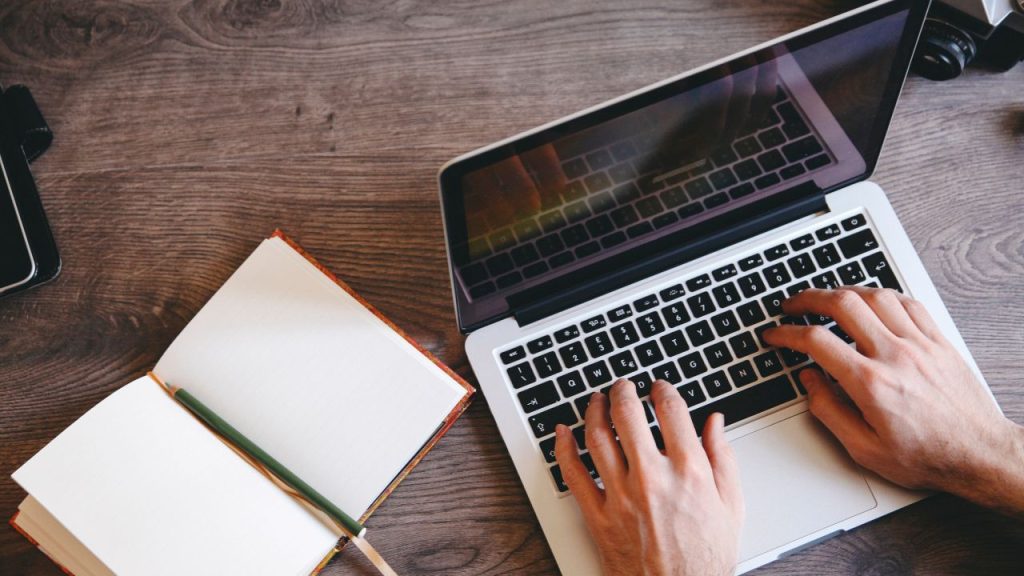 Two light-skinned hands typing on a MacBook on a wooden desk with a blank open notebook and pencil lying beside them.