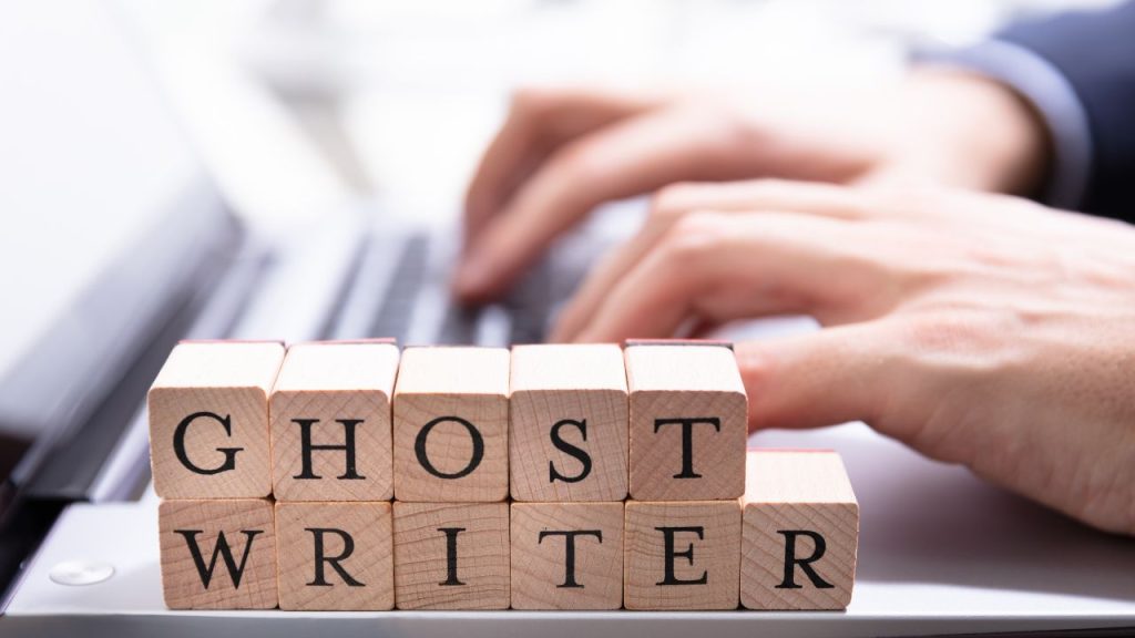 Two light-skinned hands typing on a laptop, out of focus; in the foreground, wooden blocks stacked spelling out "GHOSTWRITER" in two rows.