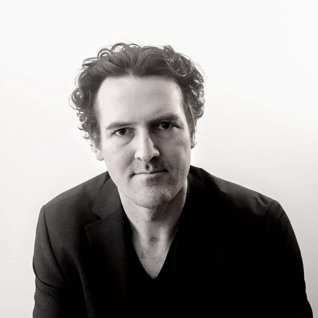 Jason Kenny, a light-skinned man with short, curly hair and a dark shirt, looks at the viewer against a plain background; the image is black and white.